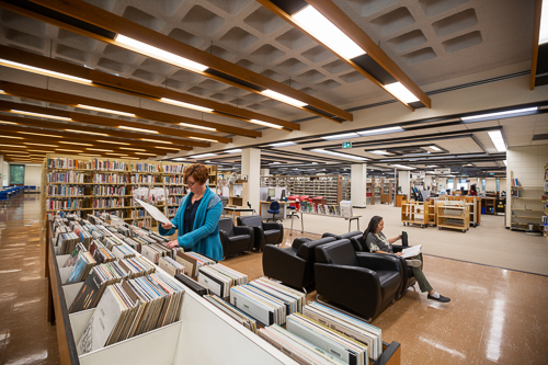A person looking at records and a second seated person reading and drinking coffee inside the Education and Music Library.